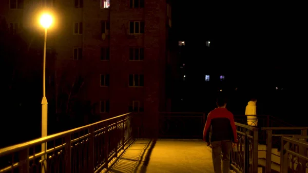 Puente peatonal con una lámpara de calle solitaria y dos hombres cruzándola por la noche. Imágenes de archivo. Vista trasera de un joven en el cruce del puente en la ciudad por la noche . — Foto de Stock