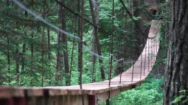 Kırmızı sırt çantalı bir gezginle yeşil sık ormandaki yürüyüş parkurunda asma köprü. Stok görüntüleri. Asılı köprüden geçen bir adamın dikiz görüntüsü.