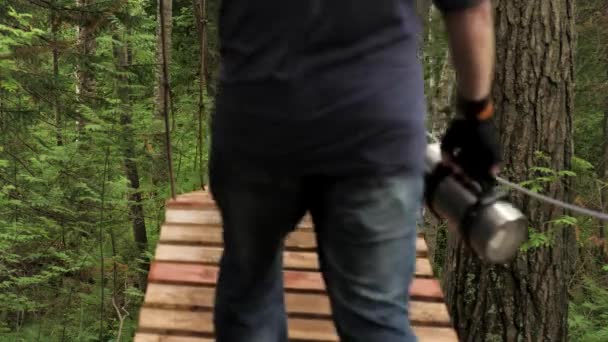 在中美洲的绿色热带丛林中远足。 库存录像。 一个手拿着金属保温瓶的人穿过被绿色森林环绕的木制吊桥的后视镜. — 图库视频影像