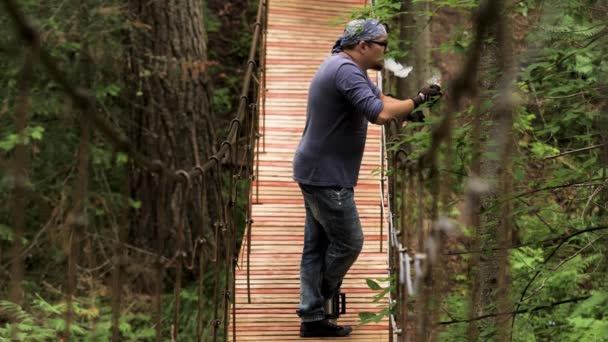 Vista lateral de un hombre parado en el puente colgante de madera y fumando una pipa mientras observa a lo lejos. Imágenes de archivo. Senderista descansando y disfrutando del bosque de verano . — Vídeos de Stock