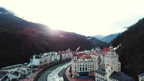 背景には明るい夕日と山のふもとの建物、ケーブルカー、川の写真。ストック映像だ山間部の小さな町や川の上を飛ぶ. — ストック動画