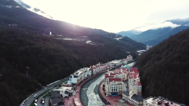 Fotos von Gebäuden, Seilbahn, Fluss am Fuße der Berge mit dem hellen Sonnenuntergang im Hintergrund. Archivmaterial. fliegen über der bergigen Kleinstadt und einem Fluss. — Stockvideo