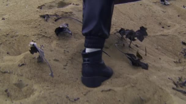 Κοντινό πλάνο μιας γυναίκας με φθινοπωρινές μπότες που περπατάει στην άμμο. Στικ. Όμορφη νεαρή γυναίκα περπατά κατά μήκος της παραλίας άμμο σε συννεφιασμένη ημέρα του φθινοπώρου — Αρχείο Βίντεο