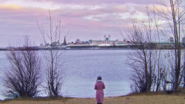 Vrouw wandelen op het strand op de achtergrond van de oude stad in de herfst. Voorraadbeelden. Achteraanzicht van aantrekkelijke jonge vrouw wandelen op herfststrand op de achtergrond van water en stad met bewolkte lucht — Stockvideo