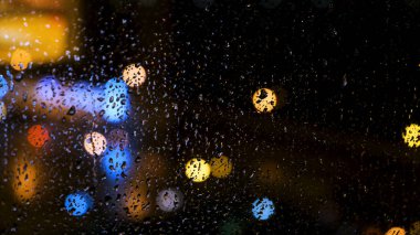 Pencerenin arkasında, üzerinde su damlaları olan büyük bir şehrin güzel odaksız parlak ışıkları. Kavram. Cam pencereye yağmur damlaları ve bulanık bokeh arkaplan.