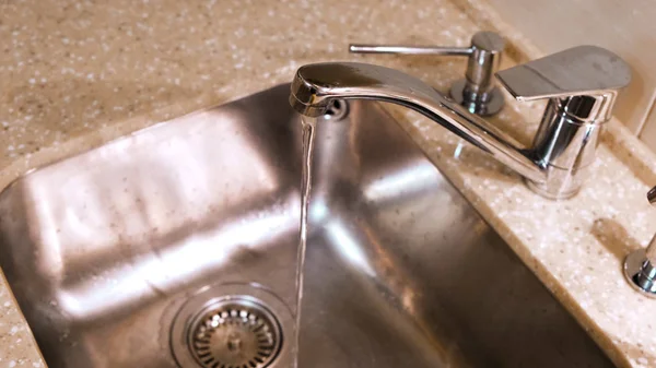 Water flows from the kitchen faucet into the metal sink. Concept. Clean water flowing from a chrome tap in the kitchen. — Stock Photo, Image