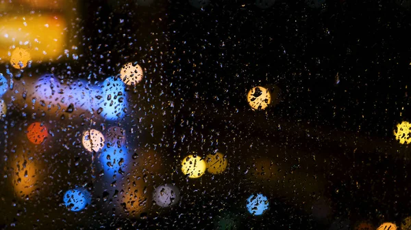 Gyönyörű elterelt fényes fények egy nagy város az ablak mögött, vízcseppekkel. Fogantatás. Eső cseppek az üvegablakra és homályos bokeh háttér. — Stock Fotó
