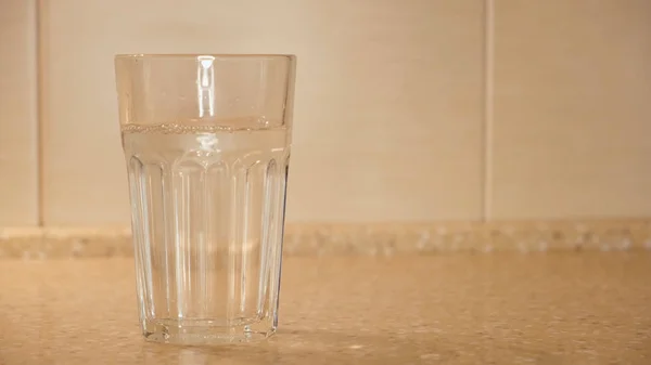 Närbild av vatten hälls i fasetterad transparent glas. Begreppet. Rent kokt vatten hälls i transparent glas på bakgrund av gamla kök kakel vägg — Stockfoto