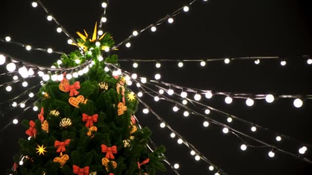 Blick von unten auf die Spitze eines schönen Weihnachtsbaums, der nachts auf der Straße mit Spielzeug und Girlanden geschmückt ist. Konzept. bunte Lichter und Bänder auf grüner Fichte auf schwarzem Himmelshintergrund. — Stockvideo