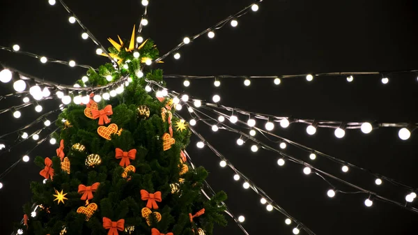 Dolní pohled na vrchol krásného vánočního stromečku zdobeného v noci hračkami a girlandami na ulici. Koncept. Barevná světla a stuhy na zeleném smrku na černém pozadí oblohy. — Stock fotografie