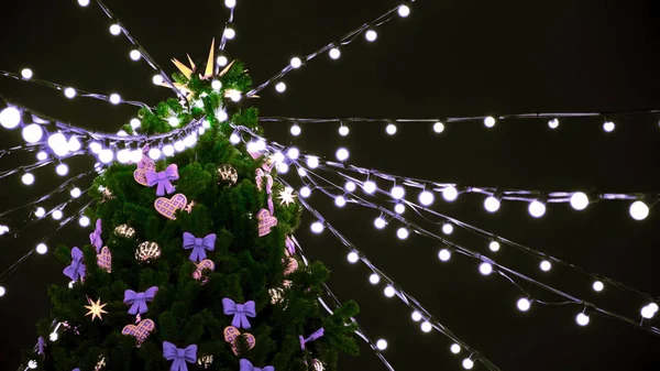 Bottom view of the top of a beautiful Christmas tree decorated with toys and garlands in the street at night. Concept. Colorful lights and ribbons on green spruce on black sky background. — Stock Photo, Image