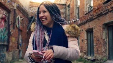 Rasta bukleli, telefonla gülümseyen zeki genç bir kadın. Stok görüntüleri. Renkli, modern görünümlü genç bir kadın telefonunda oturuyor ve eski sokakların arka planında gülüyor.