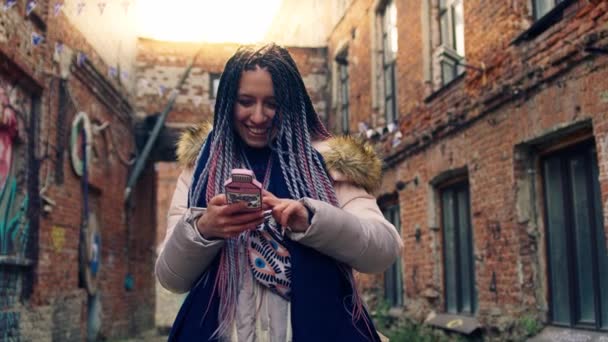 Jovem hipster sorri e se senta em seu telefone. Imagens de stock. Jovem elegante com dreadlocks coloridos com telefone no fundo de ruas antigas com casas de tijolo — Vídeo de Stock