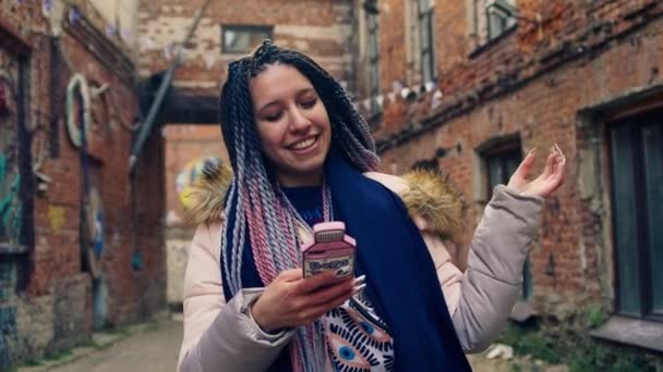 De jonge hippe vrouw glimlacht en zit op haar telefoon. Voorraadbeelden. Stijlvolle jonge vrouw met kleurrijke dreadlocks met telefoon op de achtergrond van oude straten met bakstenen huizen — Stockvideo