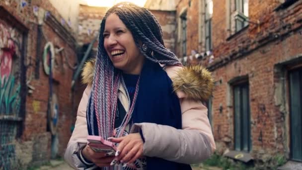 Heldere jonge vrouw met dreadlocks glimlacht met telefoon. Voorraadbeelden. Jonge vrouw in kleurrijke moderne afbeelding zit op haar telefoon en lacht op de achtergrond van oude straten — Stockvideo