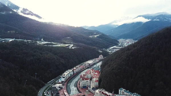 Aerial of the hotel complex situated in mountains covered by morning fog. Stock footage. Flying over green autumn forest near snowy mountain peaks.