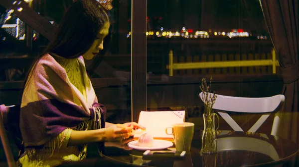 Женщина ест десерт в кафе на фоне города. Запись. Красивая молодая женщина ест десерт и читает книгу в ресторане на фоне ночного города — стоковое фото