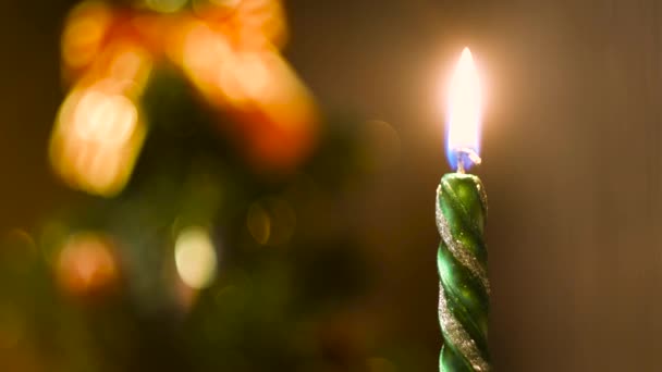 Brennende Kerze auf einem verschwommenen bunten weihnachtlichen Hintergrund. Konzept. Nahaufnahme der Flamme der grünen Neujahrskerze vor einer defokussierten Fichte. — Stockvideo