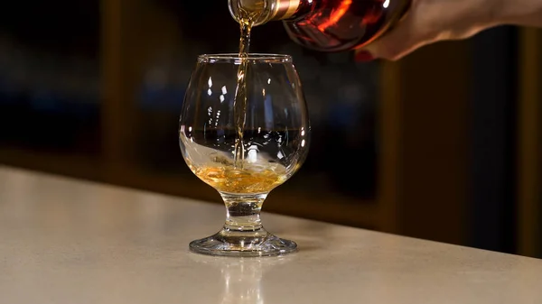Primo piano di donna mano versando cognac al vetro in piedi sul bancone del bar. Filmati delle scorte. Mano versando alcol in un bicchiere sullo sfondo sfocato di una stanza buia . — Foto Stock