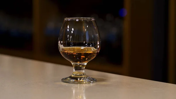 Uzavřete sklenici s koňakem, brandy nebo whisky stojící na barovém pultu. Záběry ze skladu. Silný silný alkoholický nápoj uvnitř sklenice na rozmazaném pozadí tmavé místnosti. — Stock fotografie