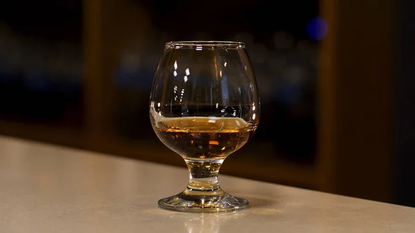 Close up of a glass with cognac, brandy or whisky standing on the bar counter. Stock footage. Strong strong alcoholic beverage inside a glass on the blurred background of a dark room. — Stock Photo, Image