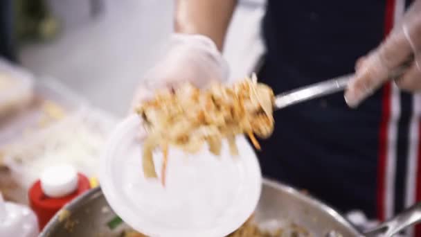 Primer plano de manos de cocinero poniendo fideos asiáticos en el plato desechable, concepto de comida callejera asiática. Art. Chef en guantes de cocina preparando comida al aire libre . — Vídeo de stock