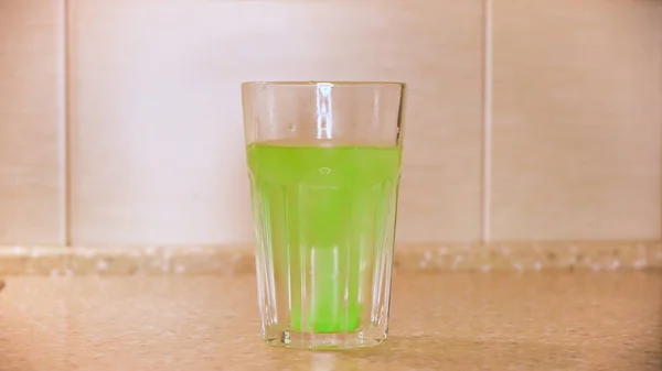 Nahaufnahme einer grünen Tablette zur Zubereitung von hausgemachtem Mineralwasser in einem Glas Wasser, das auf dem Tisch an der Wand steht. Konzept. kohlensäurehaltiges Fruchtgetränk. — Stockfoto