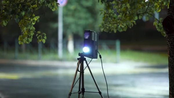 Projetor luzes feixe para mostrar foto e vídeo materiais na rua à noite no fundo da estrada vazia. Arte. Vídeo projetor e tripé ao ar livre na cidade . — Fotografia de Stock