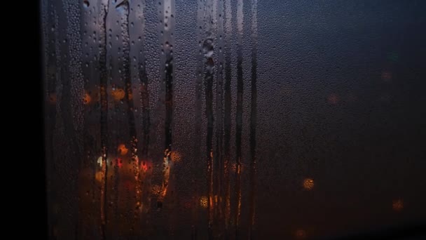 Nærme regnflekker på vindu i bakgrunnen av tåkete lys. Konsept. Regndråper som sklir nedover vinduet på bakgrunn av nattlys i by og bil – stockvideo