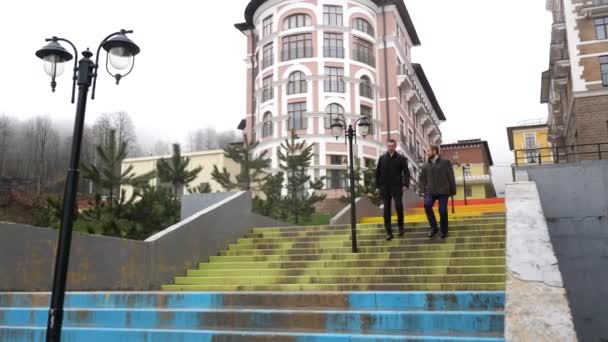 Empresarios exitosos bajando las escaleras con tiempo lluvioso cerca de un hermoso edificio hispánico y un bosque otoñal. Imágenes de archivo. Dos hombres caminando y hablando entre sí . — Vídeos de Stock