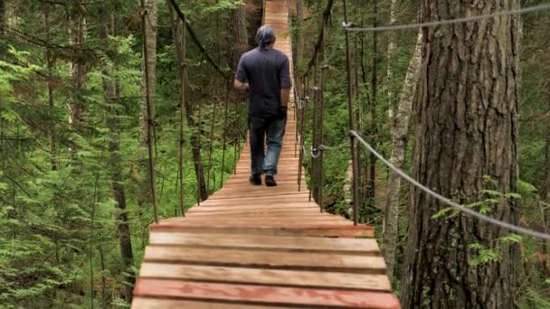 Rear view of male tourist crossing wooden suspension bridge in green forest. Stock footage. Back view of a man walking on the wooden bridge among summer green trees, active lifestyle concept. — Stok video