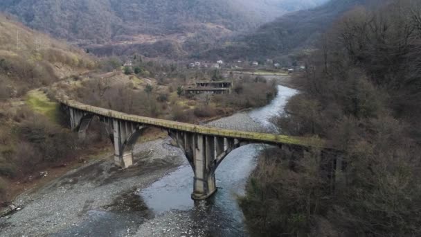 Aerial of small village situated in the valley near high forested mountains. Shot. Flying over the old concrete bridge covered by green moss and narrown cold river. — Stock Video