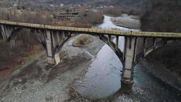 Old abandoned stone bridge over the cold mountainous river. Shot. Flying over rusty bridge and destroyed buildings amont autumn trees. — Stock Video