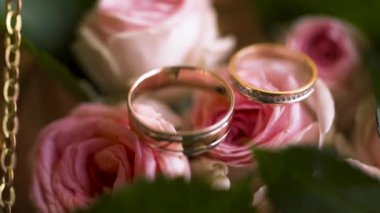 Close up of a pair of wedding rings in a decorated box with pink tender rose flowers, love and family concept. Action. Two golden yellow wedding rings lying on pink rose buds with green petals.