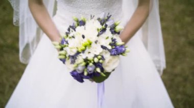 Close up of vivid wedding bouquet at young bride on green grass background, family and celebration concept. Action. Slim woman in white dress holding beautiful wedding bouquet of purple and white