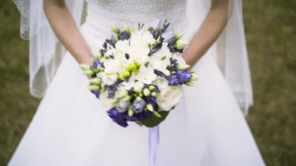 Close up of vivid wedding bouquet at young bride on green grass background, family and celebration concept. Action. Slim woman in white dress holding beautiful wedding bouquet of purple and white — 图库视频影像
