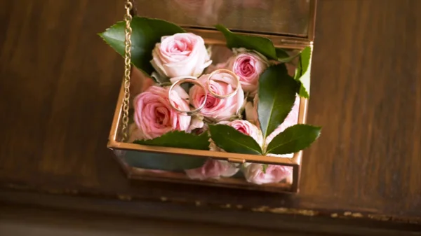 Close up of a pair of wedding rings in a decorated box with pink tender rose flowers, love and family concept. Action. Two golden yellow wedding rings in a glass box on wooden table background. — Stock Photo, Image