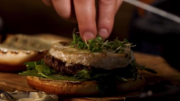 Close up of male hands putting green microgreen of mustard on camembert cheese of homemade hamburger with sauce, foodporn concept. Stock footage. Preparation of fat tasty burger. — Αρχείο Βίντεο