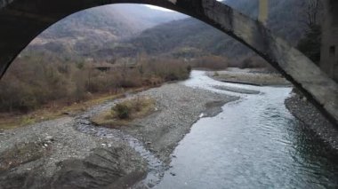Flying under the destroyed, broken concrete bridge against the background of autumn forest and a river with stony shore. Shot. Aerial of cold mountainous stream and forested hills.