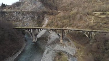 An ancient stone bridge crossing the narrow river with stony shore. Shot. Aerial of forested hills and steep cliffs near the mountainous stream.