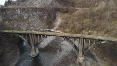 An ancient stone bridge crossing the narrow river with stony shore. Shot. Aerial of forested hills and steep cliffs near the mountainous stream.