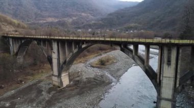 Aerial of abandoned concrete bridge covered by moss over the narrow river. Shot. Autumn landscape with high forested mountains and the small village.