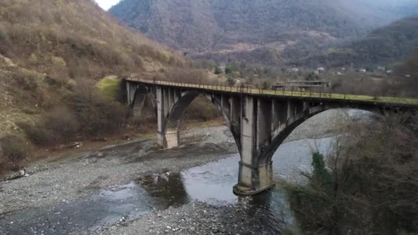 Aerial of abandoned concrete bridge covered by moss over the narrow river. Shot. Autumn landscape with high forested mountains and the small village. — Stok video