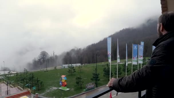 Paesaggio piovoso e nebbioso con alberi e nebbia su tutte le colline boscose e il cortile di fronte all'hotel con prato verde. Filmati delle scorte. Due uomini che conversano in piedi sul balcone . — Video Stock