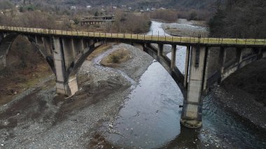 Flying under the destroyed, broken concrete bridge against the background of autumn forest and a river with stony shore. Shot. Aerial of cold mountainous stream and forested hills.
