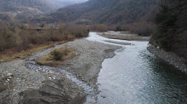 Flying under the destroyed, broken concrete bridge against the background of autumn forest and a river with stony shore. Shot. Aerial of cold mountainous stream and forested hills.
