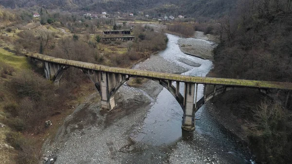 Voando sob a ponte de concreto destruída e quebrada contra o fundo da floresta de outono e um rio com costa pedregosa. Atingido. Ar de corrente montanhosa fria e colinas arborizadas . — Fotografia de Stock