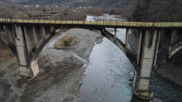 Ponte de pedra abandonada velha sobre o rio montanhoso frio. Atingido. Voando sobre ponte enferrujada e edifícios destruídos entre árvores de outono . — Fotografia de Stock