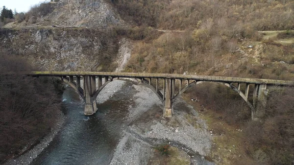 An ancient stone bridge crossing the narrow river with stony shore. Shot. Aerial of forested hills and steep cliffs near the mountainous stream. — Stockfoto