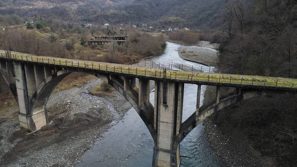 Old abandoned stone bridge over the cold mountainous river. Shot. Flying over rusty bridge and destroyed buildings amont autumn trees. — Stock Photo, Image